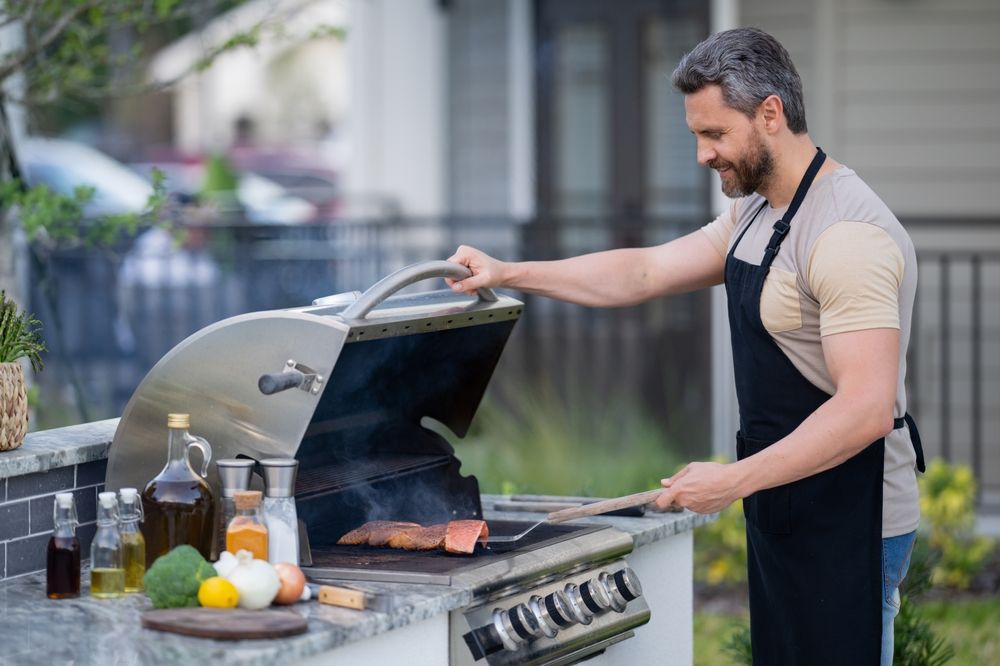 A man uses an outdoor grill to heat up steak. He is wearing a black apron and using a spatula to flip it