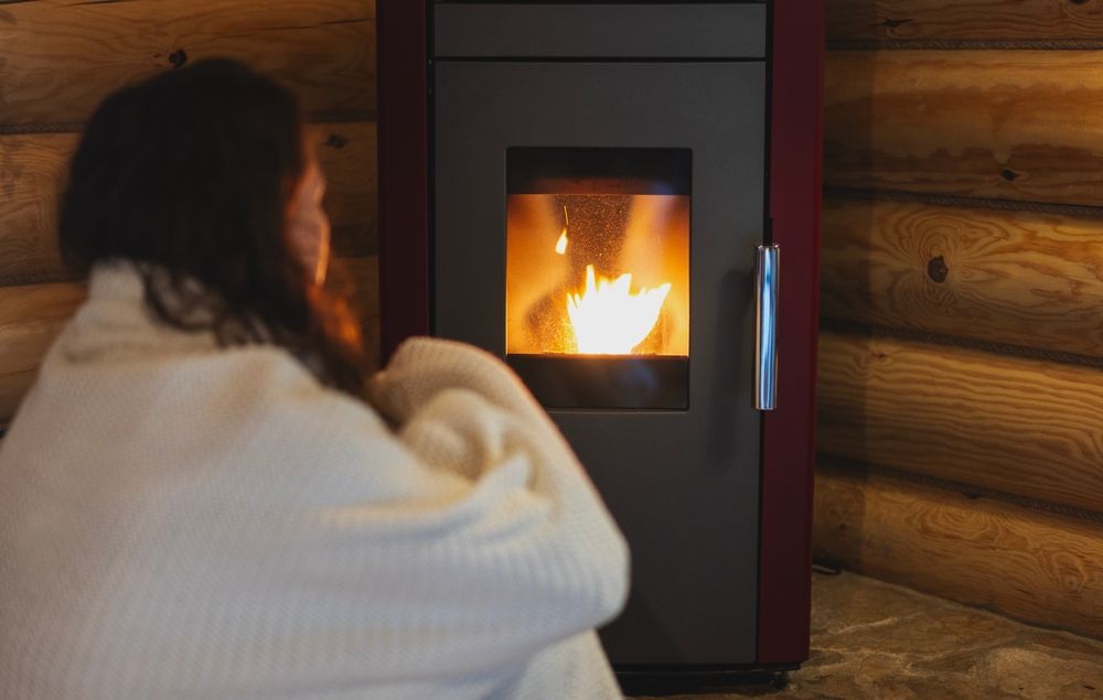 A woman wrapped in a blanket sits by a pellet stove emanating a fire